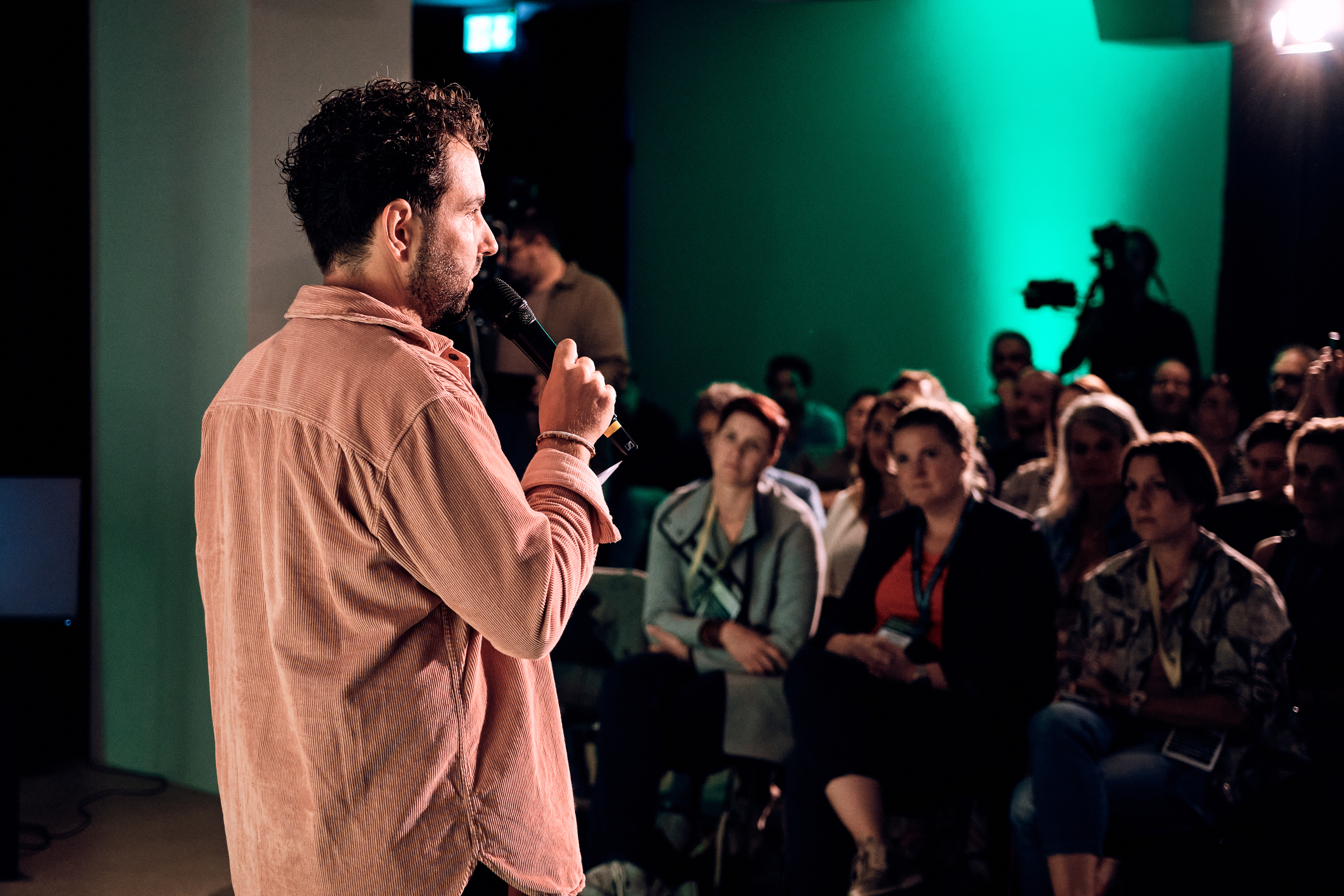 Jemand spricht in ein Mikrofon vor einem sitzenden Publikum bei einer Indoor-Veranstaltung, wobei der Hintergrund grün beleuchtet ist.